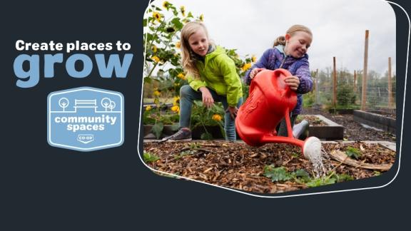 A child waters an outdoor garden with a large red watering can while her friend looks on. Wording reads 'Create place to grow' with the Community Spaces logo below.